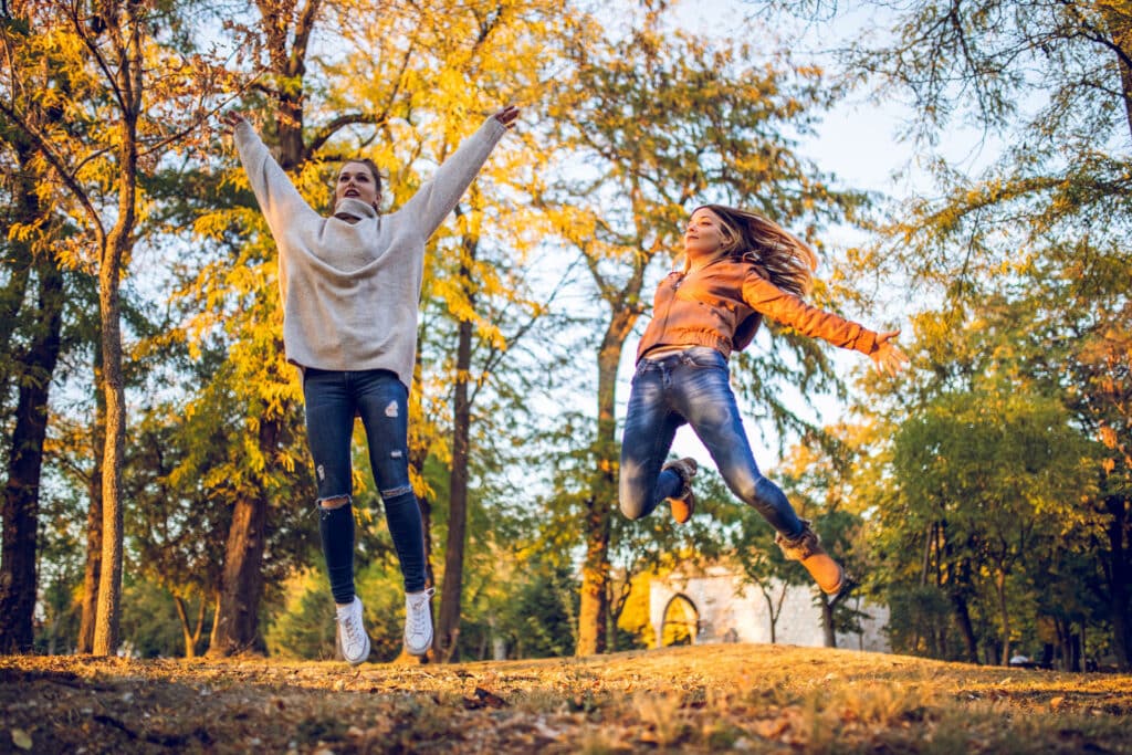 Two young beautiful caucasian women jumping in public park surrounded with autumn leafs.