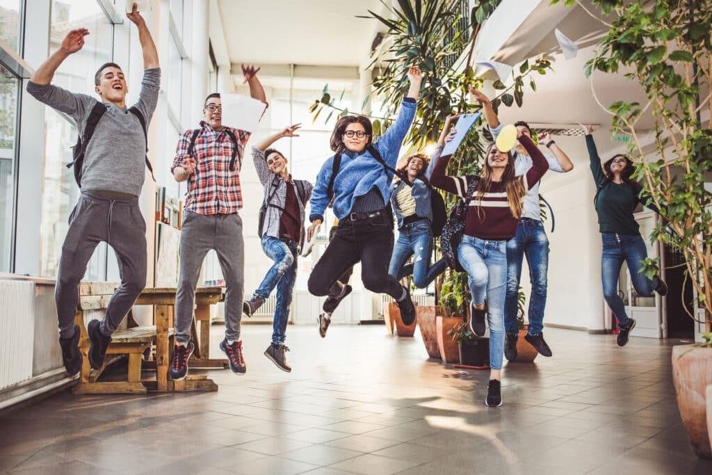 Large group of happy high school students having fun while jumping with raised arms in a hallway. Copy space.
