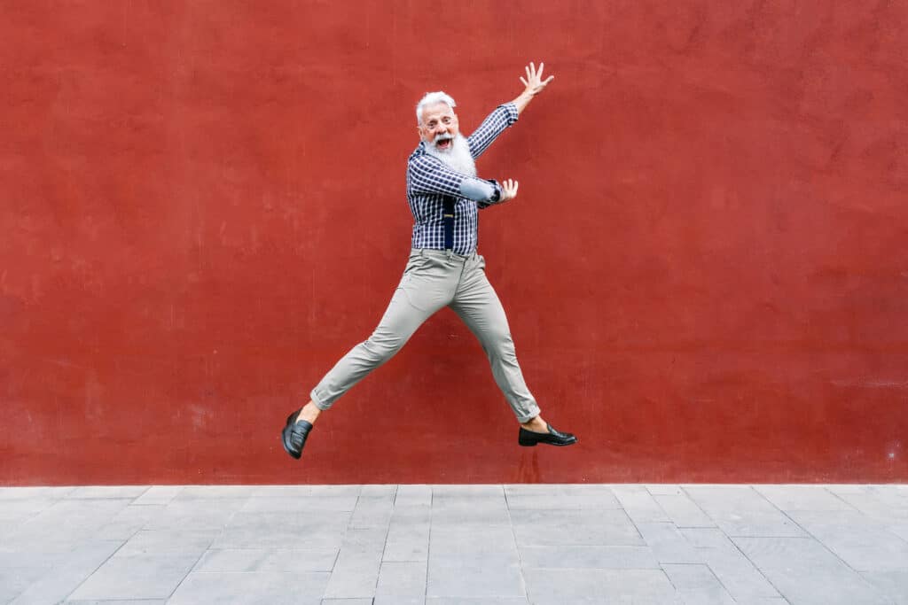 Handsome well dressed Caucasian senior man with gray beard and hair jumping and smiling with red wall behind him.