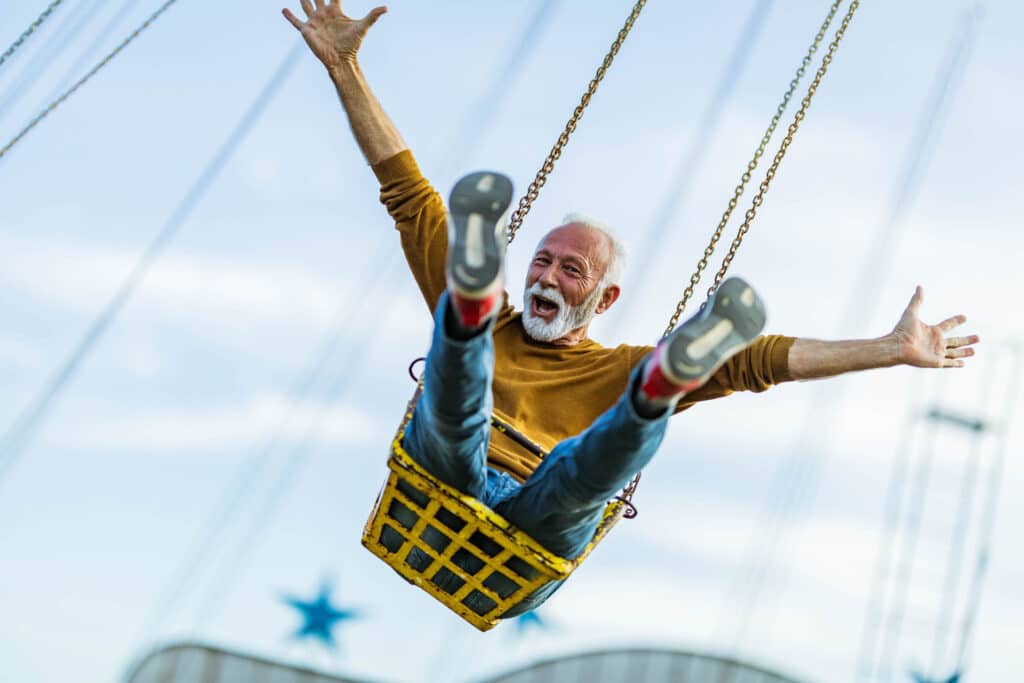 Happy senior man having fun while riding on chain swing at amusement park and looking at camera.