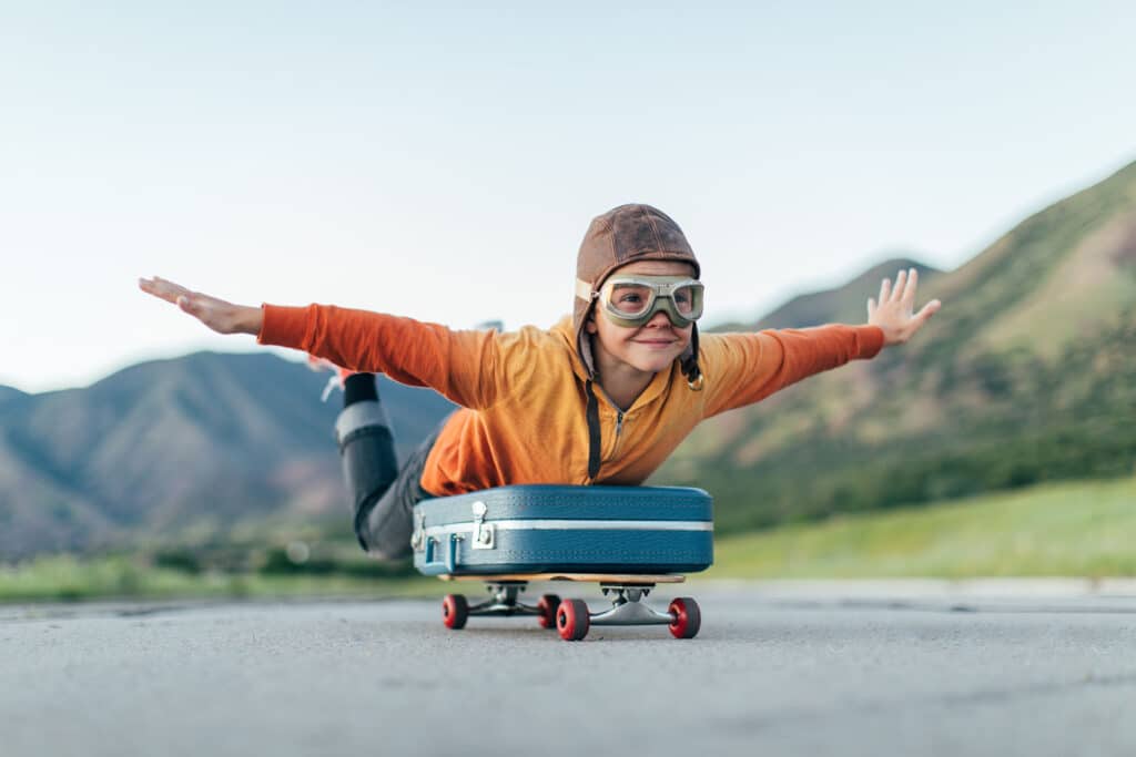 A young boy wearing flying goggles and flight cap has packed his suitcase and has placed it on a skateboard. He is ready to fly away with arms outstretched to the destination of his dreams. Image taken in Utah, USA.