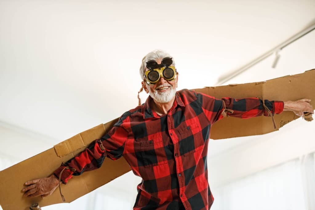 Low angle view of playful senior man having fun while pretending to fly with goggles at home. Copy space.