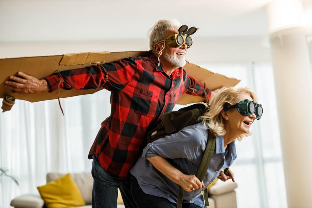 Playful senior couple having fun while pretending to fly with goggles at home. Focus is on man.