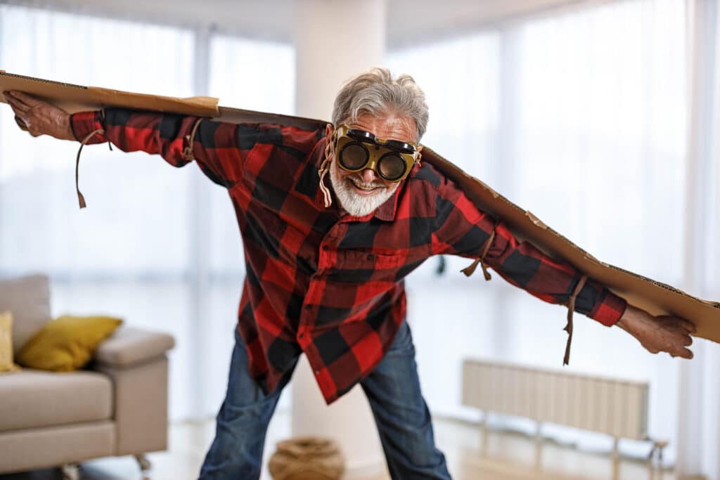 Playful senior man having fun while pretending to fly with goggles at home.